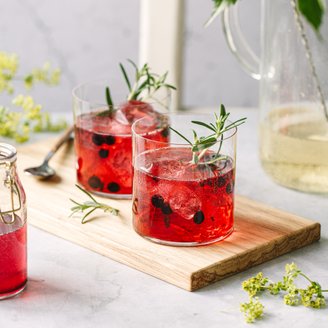 Zwei Gläser mit rotem Getränk, gefüllt mit Eis und Beeren, stehen auf einem Holzbrett neben einer kleinen Glasflasche.