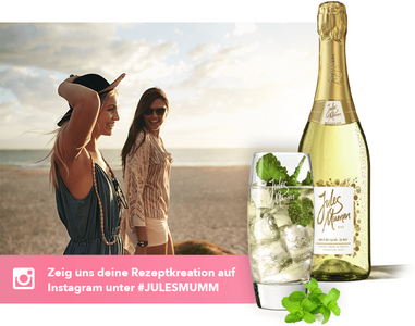 Im Vordergrund steht ein Glas mit einem erfrischenden Getränk, während im Hintergrund zwei Freundinnen am Strand flanieren.