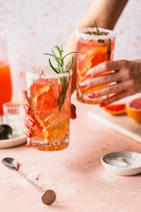 Zwei Gläser mit einem fruchtigen Getränk, garniert mit Rosmarin und Zitrusfrüchten, werden auf einem rosa Tisch serviert.