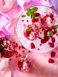 Drei Gläser mit gefrorenen Himbeeren und Granatapfelkernen auf Eis, umgeben von buntem Krepppapier.