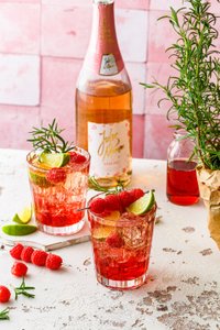 Fruchtige Cocktails mit Himbeeren und Limetten, serviert in eleganten Gläsern, umgeben von frischen Kräutern und einer Sektflasche.