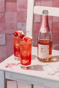 Auf einem weißen Tisch stehen zwei Cocktails mit Eis, dekoriert mit Kirschen und Grapefruit, neben einer Flasche Rosé.