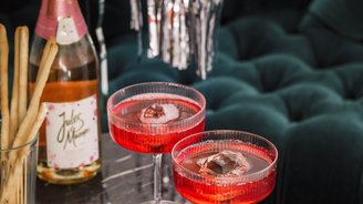 Zwei elegante Cocktails in roten Gläsern stehen auf einem marmorierten Tisch, umgeben von Snacks und einer Flasche Sekt.