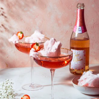 Zwei elegante Cocktails mit rosa Zuckerwatte und frischen Erdbeeren stehen auf einem marmorierten Tisch vor einer zarten rosa Wand.