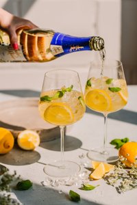 Eine Hand gießt ein sprudelndes Getränk in ein Glas, während frische Zitronen und Minzblätter im Vordergrund liegen.
