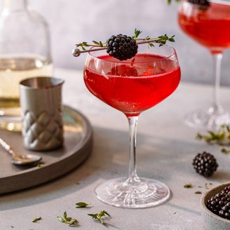 Ein eleganter Cocktail in einem Glas, garniert mit einer Brombeere und Thymian, auf einem hellen Tisch serviert.