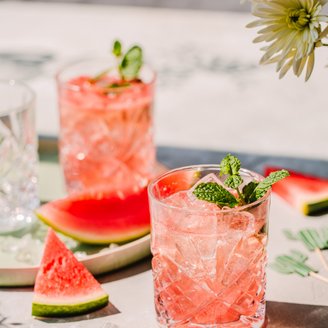 Zwei Gläser mit einem erfrischenden Wassermelonen-Cocktail, garniert mit frischer Minze, auf einem hellen Tisch.