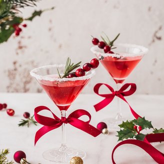 Zwei stilvolle Cocktails mit roten Schleifen, umgeben von festlichen Ornamenten und Tannenzweigen, strahlen Weihnachtsstimmung aus.