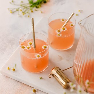 Zwei Gläser mit einem sprudelnden, rosafarbenen Getränk, dekoriert mit kleinen Kamillenblüten und goldenen Strohhalmen.