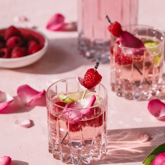Ein elegantes Glas mit einem rosa Getränk, garniert mit Himbeeren und Rosenblüten, steht auf einer zarten rosa Oberfläche.