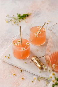 Zwei Gläser mit einem sprudelnden, rosafarbenen Getränk, dekoriert mit kleinen Kamillenblüten und goldenen Strohhalmen.