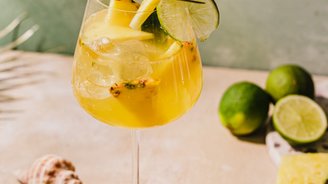 Ein erfrischender Cocktail in einem eleganten Glas, garniert mit Ananas und Limette, auf einem sandfarbenen Tisch.