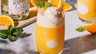 Zwei Gläser mit einem fruchtigen Cocktail, dekoriert mit einer Kugel Eis und frischen Minzblättern, auf einem hellen Tisch.