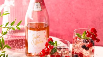 Auf einem Tisch stehen zwei Gläser mit prickelndem Rosé, dekoriert mit Beeren und Minze, während eine Flasche im Hintergrund sichtbar ist.