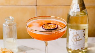 Ein leuchtend orangefarbener Cocktail mit Schaumkrone und einer Scheibe Maracuja, neben einer Flasche Jules Mumm Sekt und einem kleinen Glas.