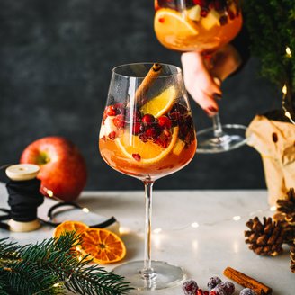 Auf einem Tisch stehen Gläser mit einem fruchtigen Getränk, umgeben von Tannenzweigen, getrockneten Orangen und Lichtern.