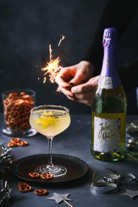 Eine Hand zündet eine Wunderkerze über einem Cocktail mit Sternenfrüchten, umgeben von Brezeln und einer Flasche Sekt.