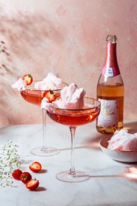Zwei elegante Cocktails mit rosa Zuckerwatte und frischen Erdbeeren stehen auf einem marmorierten Tisch vor einer zarten rosa Wand.