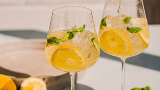 Eine Hand gießt ein sprudelndes Getränk in ein Glas, während frische Zitronen und Minzblätter im Vordergrund liegen.