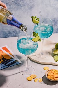 Ein Getränk mit blauer Farbe wird in ein Glas gegossen, während eine Hand die Flasche hält und frische Minze und Limette sichtbar sind.