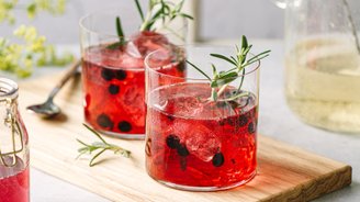 Zwei Gläser mit rotem Getränk, gefüllt mit Eis und Beeren, stehen auf einem Holzbrett neben einer kleinen Glasflasche.
