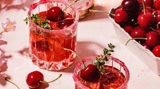 Zwei Gläser mit einem fruchtigen Cocktail, dekoriert mit Kirschen und frischen Kräutern, stehen auf einer rosa Oberfläche.