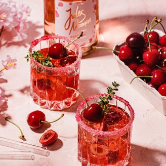 Zwei Gläser mit einem fruchtigen Cocktail, dekoriert mit Kirschen und frischen Kräutern, stehen auf einer rosa Oberfläche.