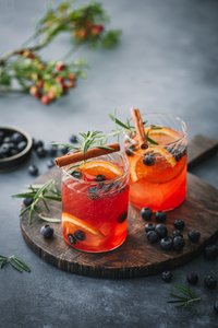 Zwei Gläser mit einem leuchtend roten Getränk, garniert mit Zimtstangen und frischen Beeren, auf einem Holzbrett.