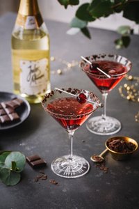 Zwei elegante Cocktails in Martinigläsern, dekoriert mit Schokoladenstückchen und einer Kirsche, stehen auf einem dunklen Tisch.
