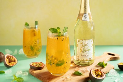 Die Gläser enthalten ein fruchtiges Getränk mit Passionsfruchtstücken und Basilikum, während die Flasche Sekt im Hintergrund leuchtet.