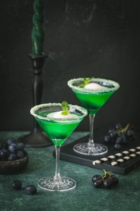 Zwei grüne Cocktails in eleganten Gläsern, dekoriert mit Minze und Zuckerrand, stehen auf einem dunklen Tisch.