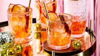 Drei Gläser mit bunten Cocktails, dekoriert mit Apfelscheiben und Zimtstangen, stehen auf einem goldenen Tisch.