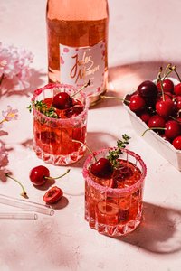 Zwei Gläser mit einem fruchtigen Cocktail, dekoriert mit Kirschen und frischen Kräutern, stehen auf einer rosa Oberfläche.