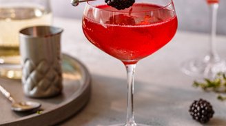 Ein eleganter Cocktail in einem Glas, garniert mit einer Brombeere und Thymian, auf einem hellen Tisch serviert.