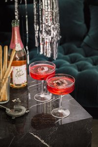 Zwei elegante Cocktails in roten Gläsern stehen auf einem marmorierten Tisch, umgeben von Snacks und einer Flasche Sekt.