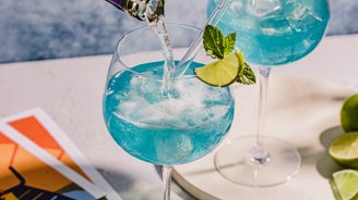 Ein Getränk mit blauer Farbe wird in ein Glas gegossen, während eine Hand die Flasche hält und frische Minze und Limette sichtbar sind.