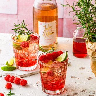 Fruchtige Cocktails mit Himbeeren und Limetten, serviert in eleganten Gläsern, umgeben von frischen Kräutern und einer Sektflasche.