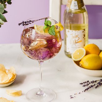 Ein elegantes Glas mit einem bunten Getränk, garniert mit Zitrone, Minze und Lavendel, steht auf einem hellen Tisch.