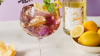Ein elegantes Glas mit einem bunten Getränk, garniert mit Zitrone, Minze und Lavendel, steht auf einem hellen Tisch.