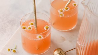 Zwei Gläser mit einem sprudelnden, rosafarbenen Getränk, dekoriert mit kleinen Kamillenblüten und goldenen Strohhalmen.