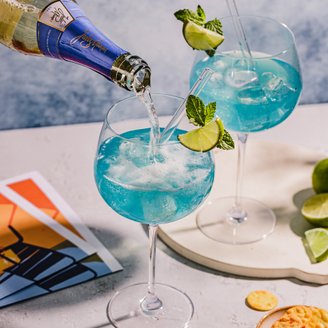 Ein Getränk mit blauer Farbe wird in ein Glas gegossen, während eine Hand die Flasche hält und frische Minze und Limette sichtbar sind.