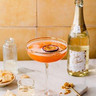 Ein leuchtend orangefarbener Cocktail mit Schaumkrone und einer Scheibe Maracuja, neben einer Flasche Jules Mumm Sekt und einem kleinen Glas.
