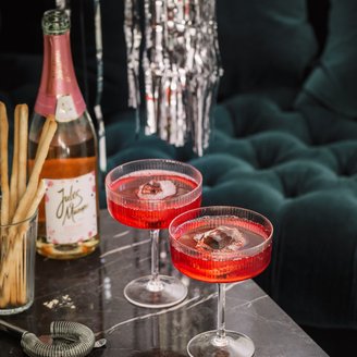 Zwei elegante Cocktails in roten Gläsern stehen auf einem marmorierten Tisch, umgeben von Snacks und einer Flasche Sekt.