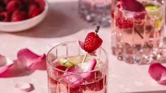 Ein elegantes Glas mit einem rosa Getränk, garniert mit Himbeeren und Rosenblüten, steht auf einer zarten rosa Oberfläche.