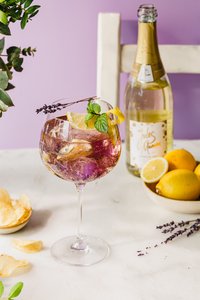 Ein elegantes Glas mit einem bunten Getränk, garniert mit Zitrone, Minze und Lavendel, steht auf einem hellen Tisch.