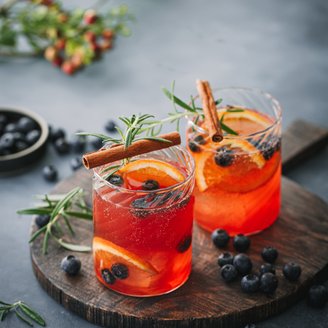 Zwei Gläser mit einem leuchtend roten Getränk, garniert mit Zimtstangen und frischen Beeren, auf einem Holzbrett.