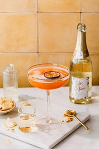 Ein leuchtend orangefarbener Cocktail mit Schaumkrone und einer Scheibe Maracuja, neben einer Flasche Jules Mumm Sekt und einem kleinen Glas.