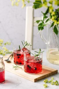 Zwei Gläser mit rotem Getränk, gefüllt mit Eis und Beeren, stehen auf einem Holzbrett neben einer kleinen Glasflasche.