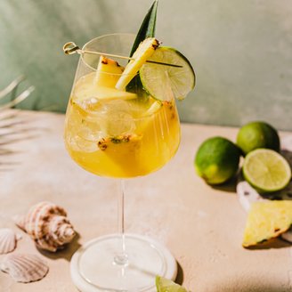 Ein erfrischender Cocktail in einem eleganten Glas, garniert mit Ananas und Limette, auf einem sandfarbenen Tisch.