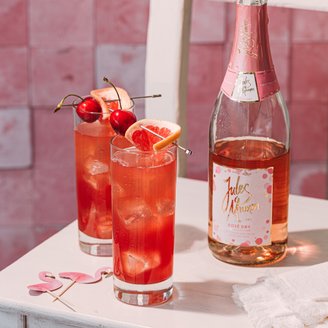 Auf einem weißen Tisch stehen zwei Cocktails mit Eis, dekoriert mit Kirschen und Grapefruit, neben einer Flasche Rosé.
